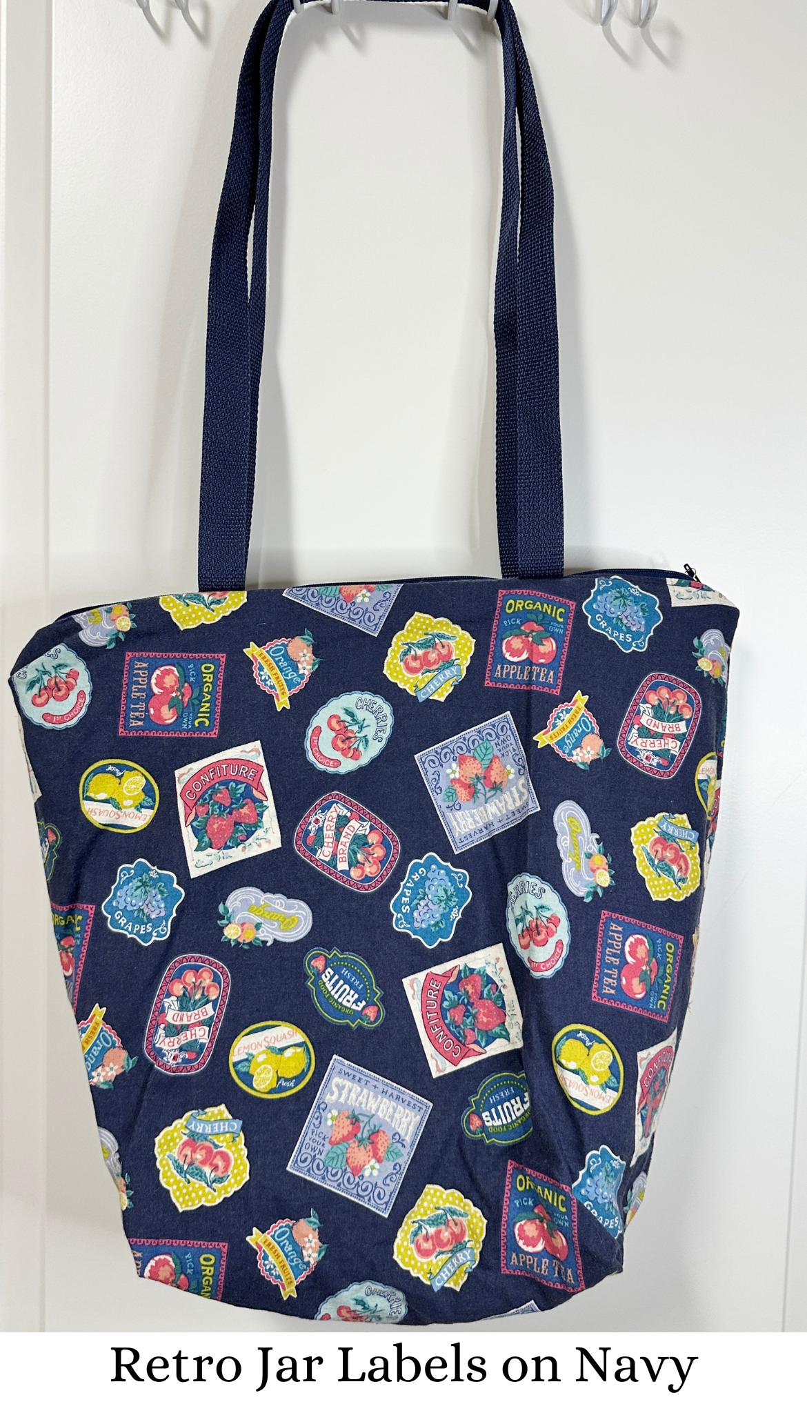 This tote has navy handles and a navy zipper; the print is various retro styled fruit and jam labels on a background of navy; the labels are bright against the background.