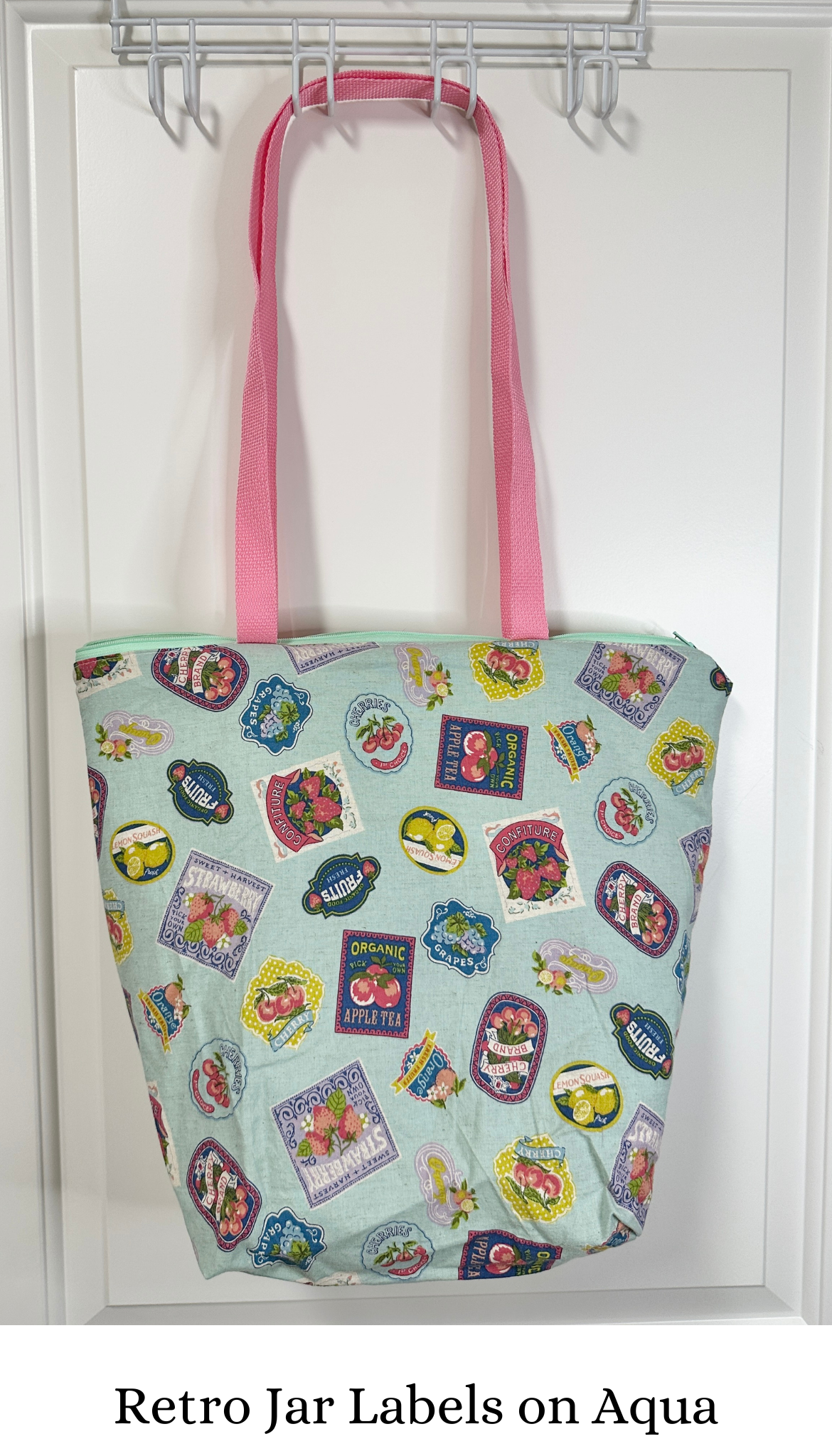 This tote has pink handles and an aqua zipper; the print is various retro styled fruit and jam labels on a background of aqua; the labels are bright against the background.