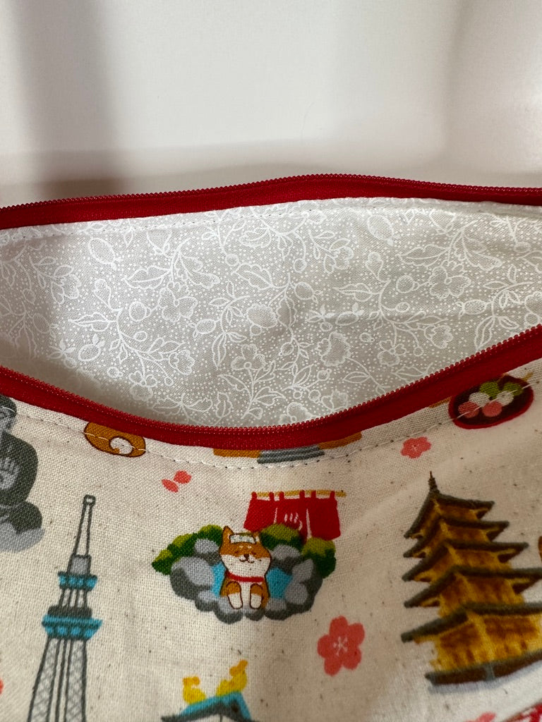 This tote has red handles and zipper. The motif is a cartoon styled Shiba Inu exploring Japan and seeing various icons like the Tokyo Tower, Sky Tree, Osaka Castle, Kinkakuji, Sensoji, and the gate at Miyajima. The background is an off-white with tiny brown speckles (reminds me of vanilla ice cream). This photo shows that the lining is the white on white floral print.