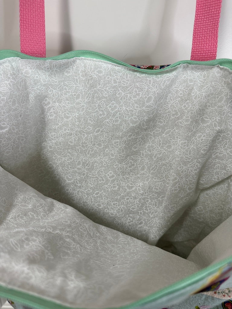 This tote has pink handles and an aqua zipper; the print is various retro styled fruit and jam labels on a background of aqua; the labels are bright against the background. This photo shows the the lining is white on white of the floral print.