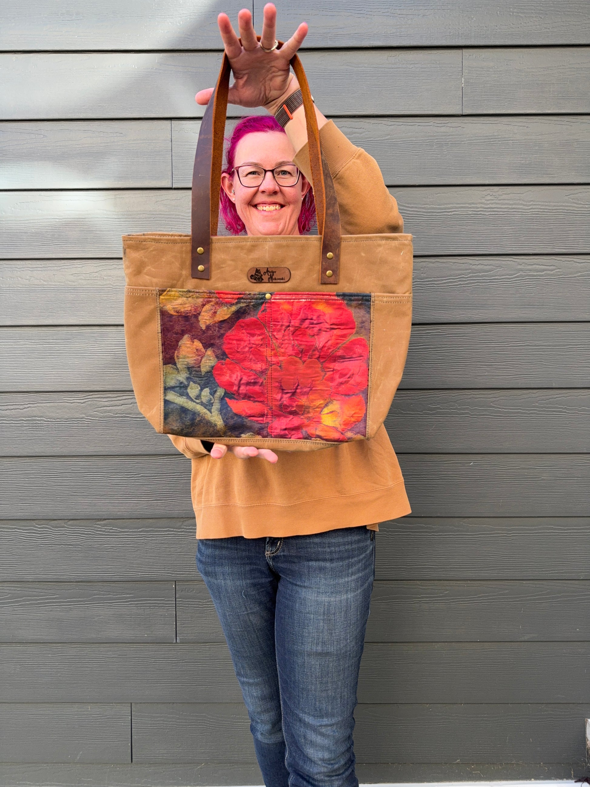 Woman with pink hair holding a tan waxed canvas bag framing a poppy design standing against a grey wall.