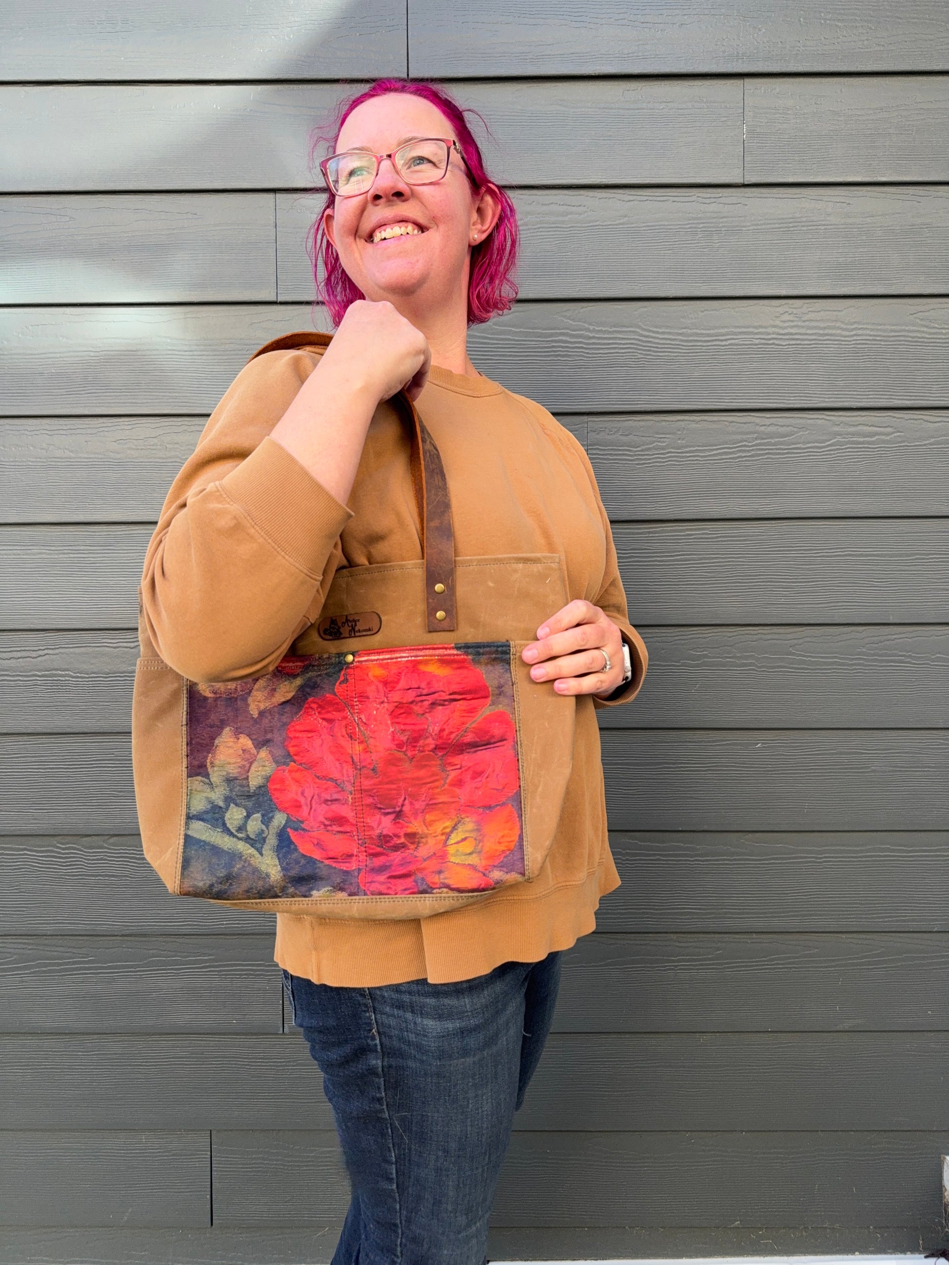 Woman holding a tote bag against a grey wall; detail is a large poppy against navy. The poppy is framed by tan waxed canvas.