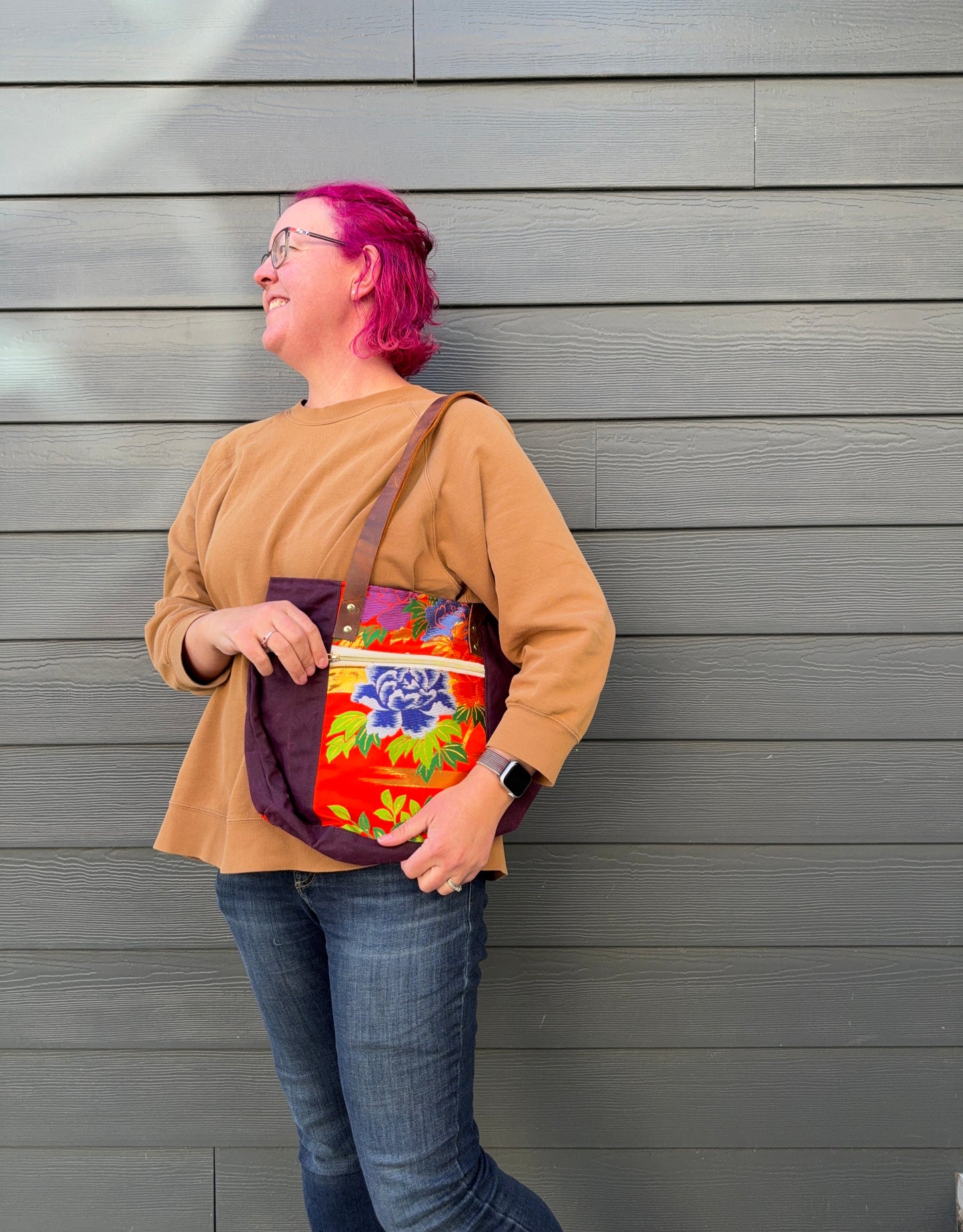 Person with pink hair holding a colorful bag against a gray wall