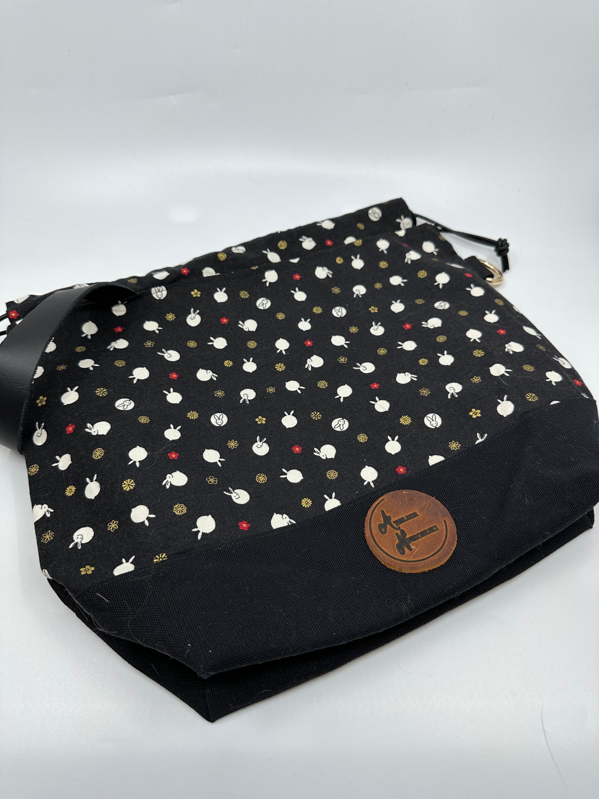 Flat lay Kato drawstring bag with black canvas bottom. The Japanese fabric upper on this bag are tiny bunnies that look like polka dots; scattered between bunny are delicate red and shimmering gold flowers. The background of the fabric is black. Drawstring, d ring tabs, and sewn-in leather handle are all black. D rings are gold.