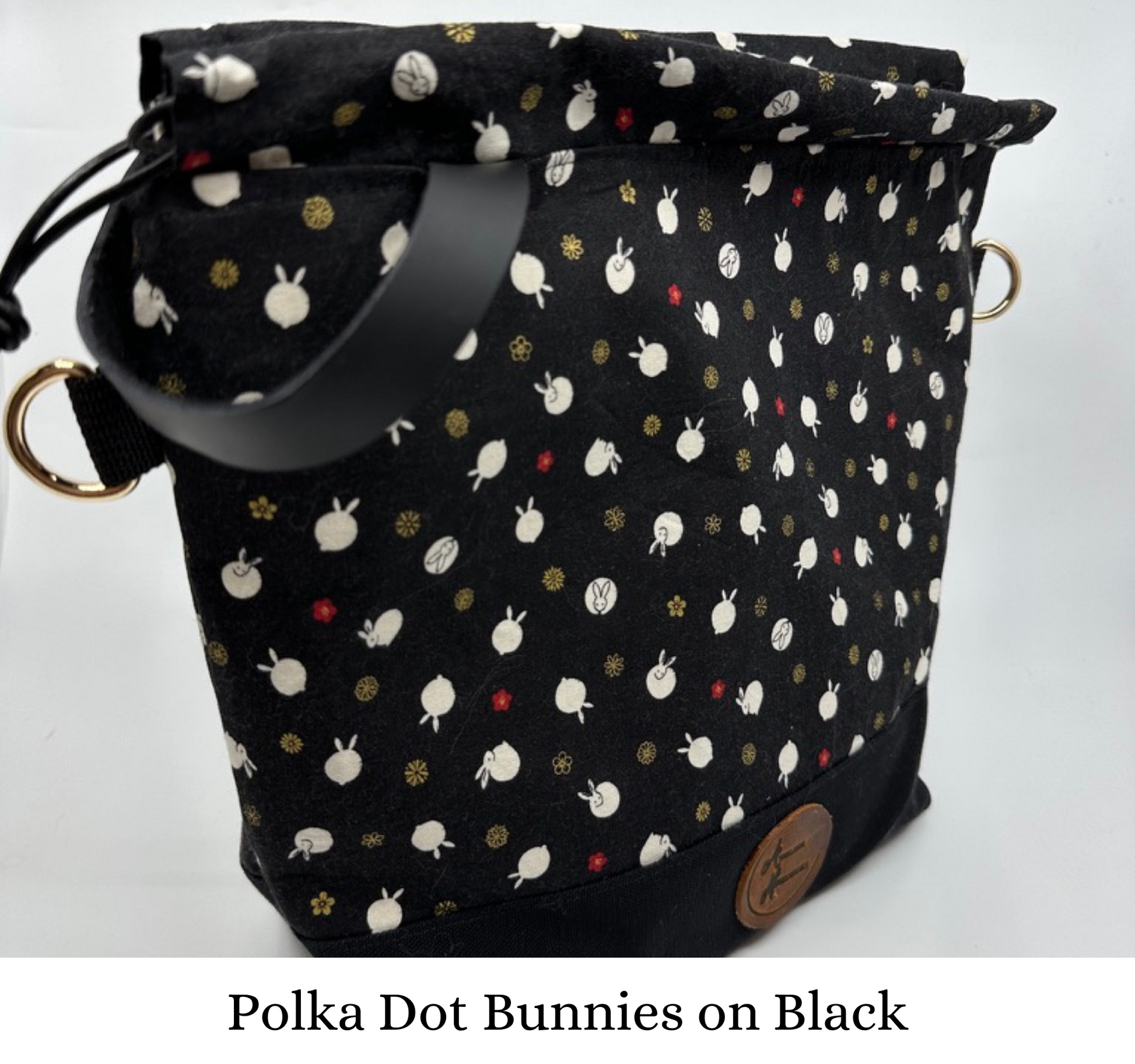 Kato drawstring bag with black canvas bottom. The Japanese fabric upper on this bag are tiny bunnies that look like polka dots; scattered between bunny are delicate red and shimmering gold flowers. The background of the fabric is black. Drawstring, d ring tabs, and sewn-in leather handle are all black. D rings are gold.