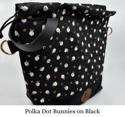 Kato drawstring bag with black canvas bottom. The Japanese fabric upper on this bag are tiny bunnies that look like polka dots; scattered between bunny are delicate red and shimmering gold flowers. The background of the fabric is black. Drawstring, d ring tabs, and sewn-in leather handle are all black. D rings are gold.