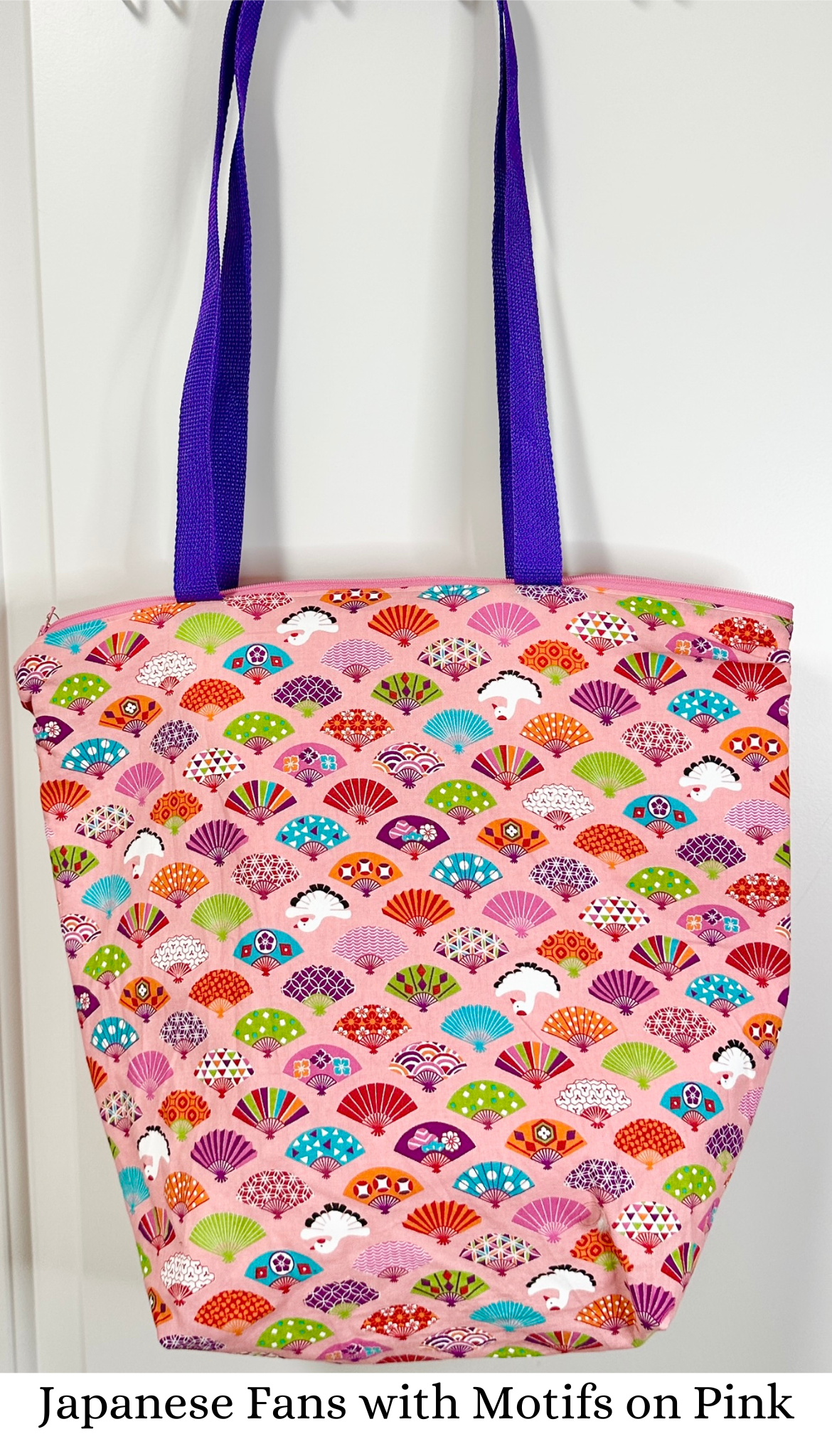 This tote bag has purple handles; the fabric print is bright fans with various Japanese styled patterns on them interspersed with white cranes, all on a bubblegum pink background.