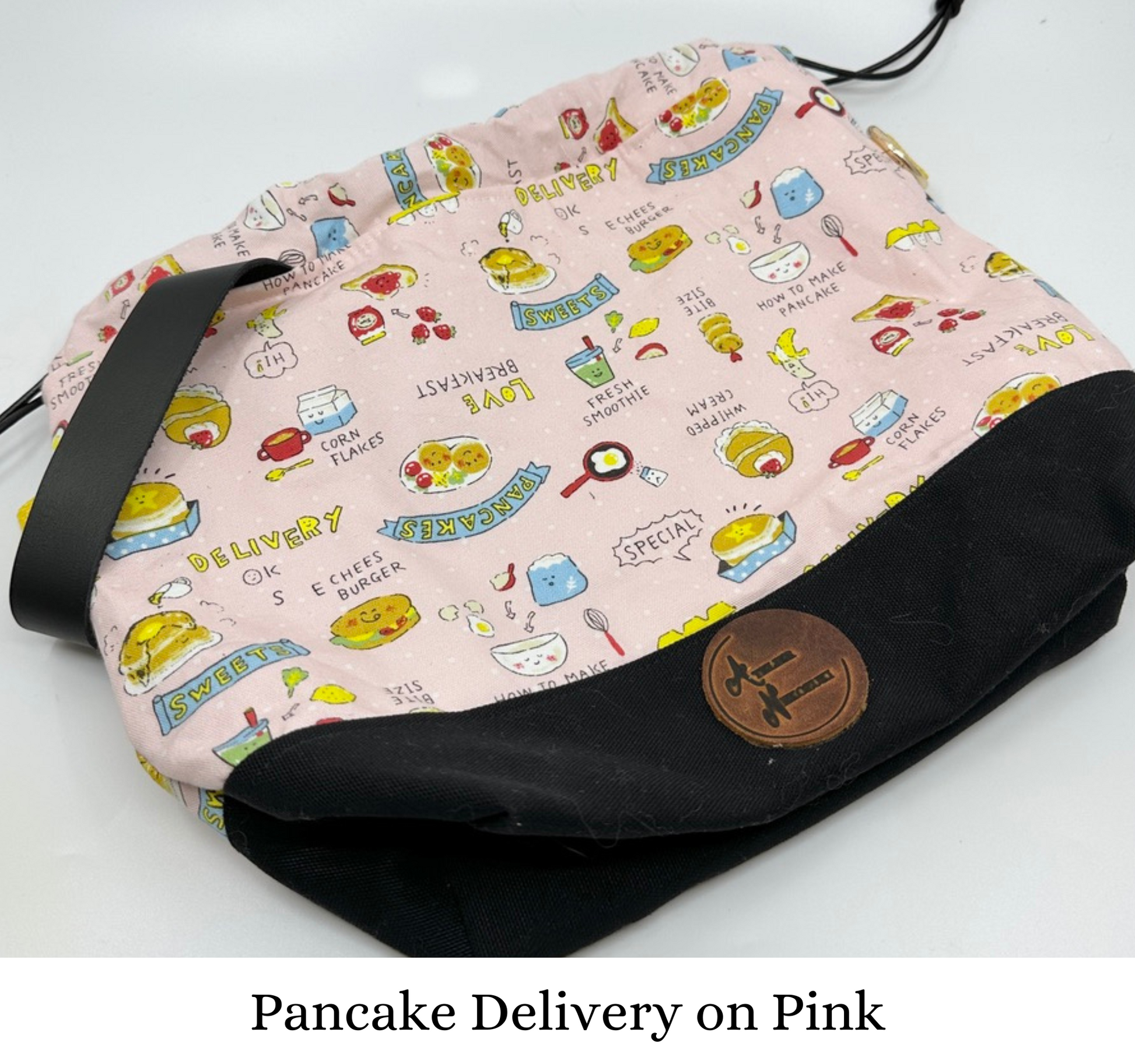 Kato drawstring bag with black canvas bottom. The Japanese fabric upper is light pink with various breakfast foods (and a random cheeseburger). Sewn in wristlet is black, drawstring is black, d ring tabs are yellow and d rings are gold.