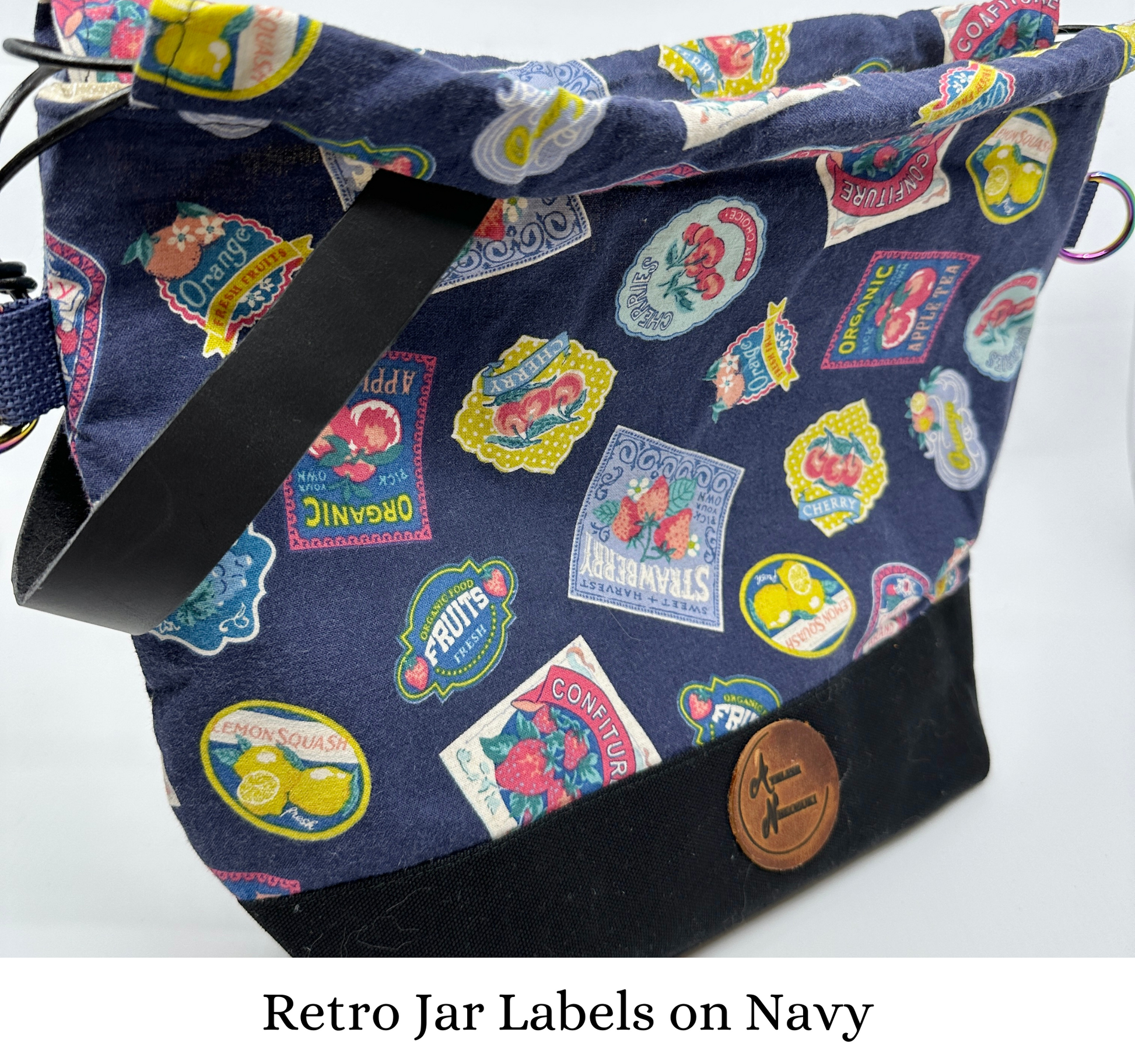 Kato drawstring bag with retro-styled jam and fruit jar labels on navy background. Canvas bottom is black; leather wristlet handle is black. D-rings for optional cross body strap are iridescent; drawstring is black leather cord.