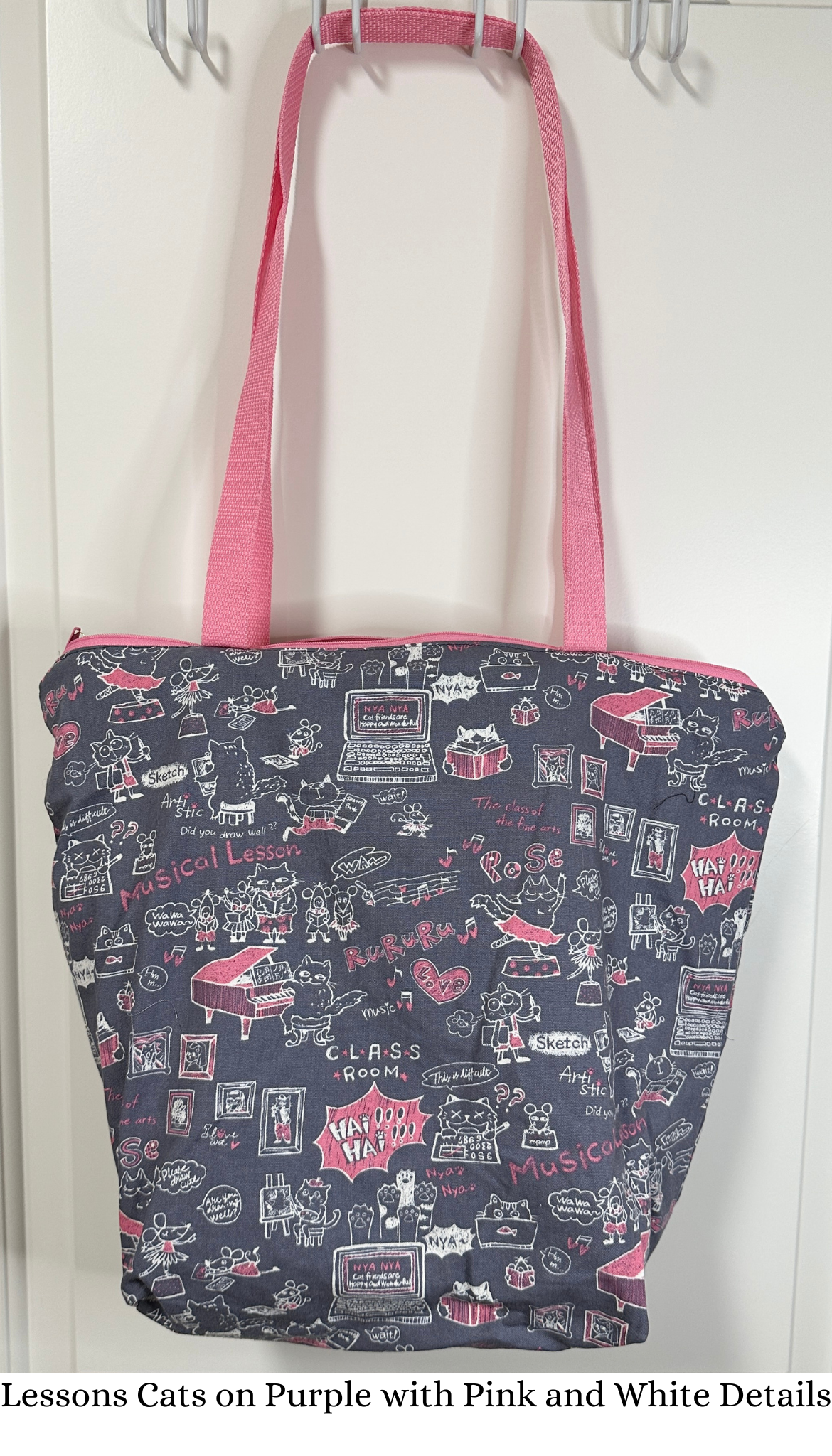 This tote bag has pink handles and a pink zipper; the fabric print is cats learning various skills (piano, etc). The cats are white and there are highlights of pink.