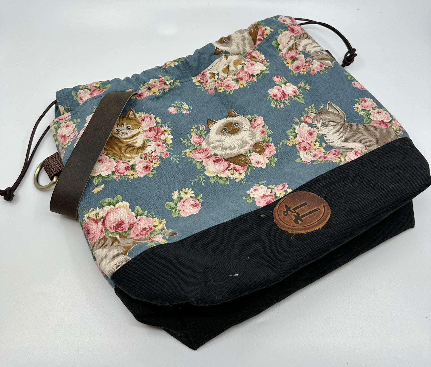 Flat lay of Kato drawstring bag with black canvas bottom. Japanese fabric upper is dusty blue with cats wreathed in florals. In the middle of the "front" of this bag is a Himalayan looking cat. The sewn in leather handle is dark brown; the drawstring is brown; the d ring tabs are brown, and the d rings are bronze. 