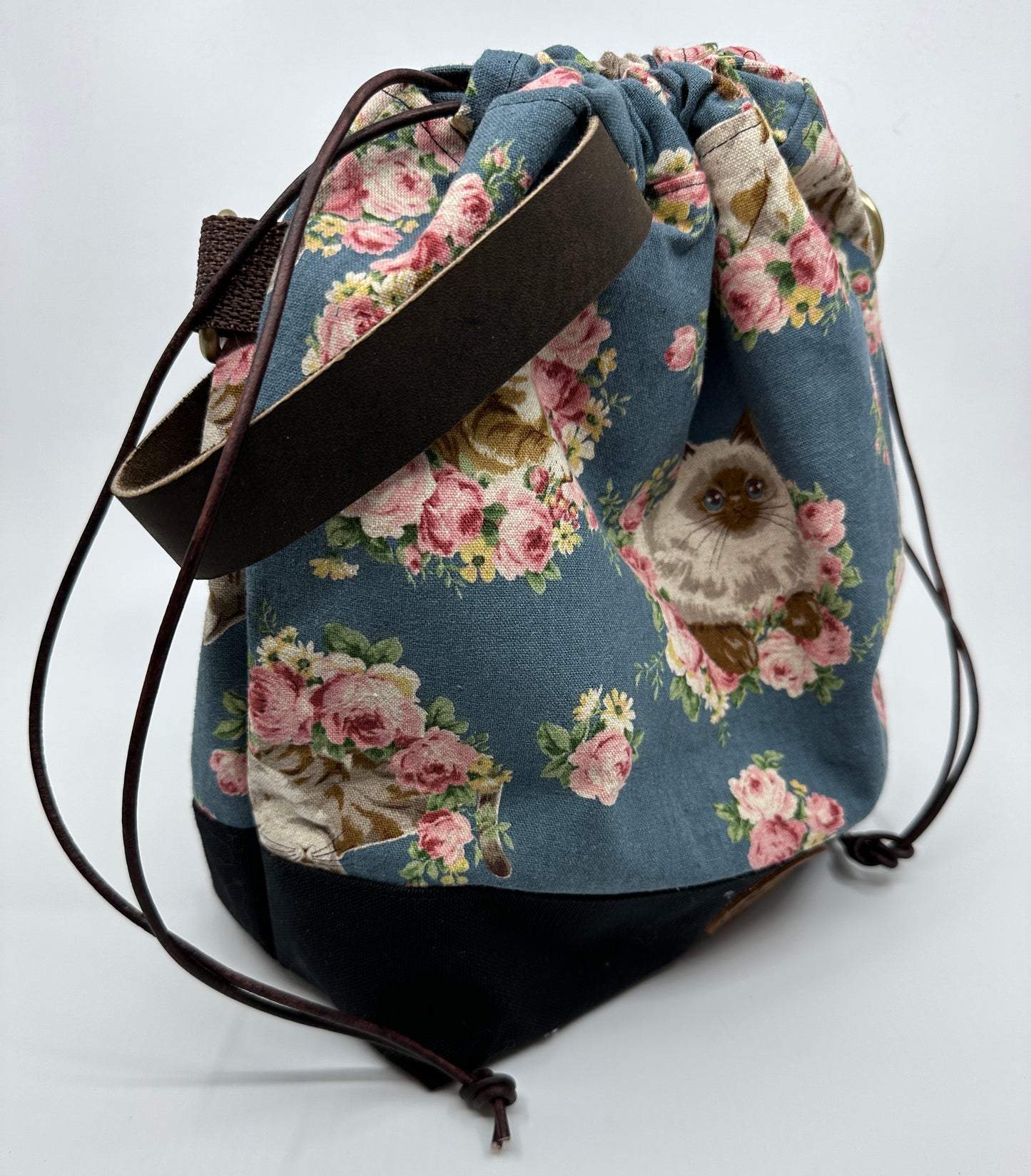 Cinched Flat lay of Kato drawstring bag with black canvas bottom. Japanese fabric upper is dusty blue with cats wreathed in florals. In the middle of the "front" of this bag is a Himalayan looking cat. The sewn in leather handle is dark brown; the drawstring is brown; the d ring tabs are brown, and the d rings are bronze.