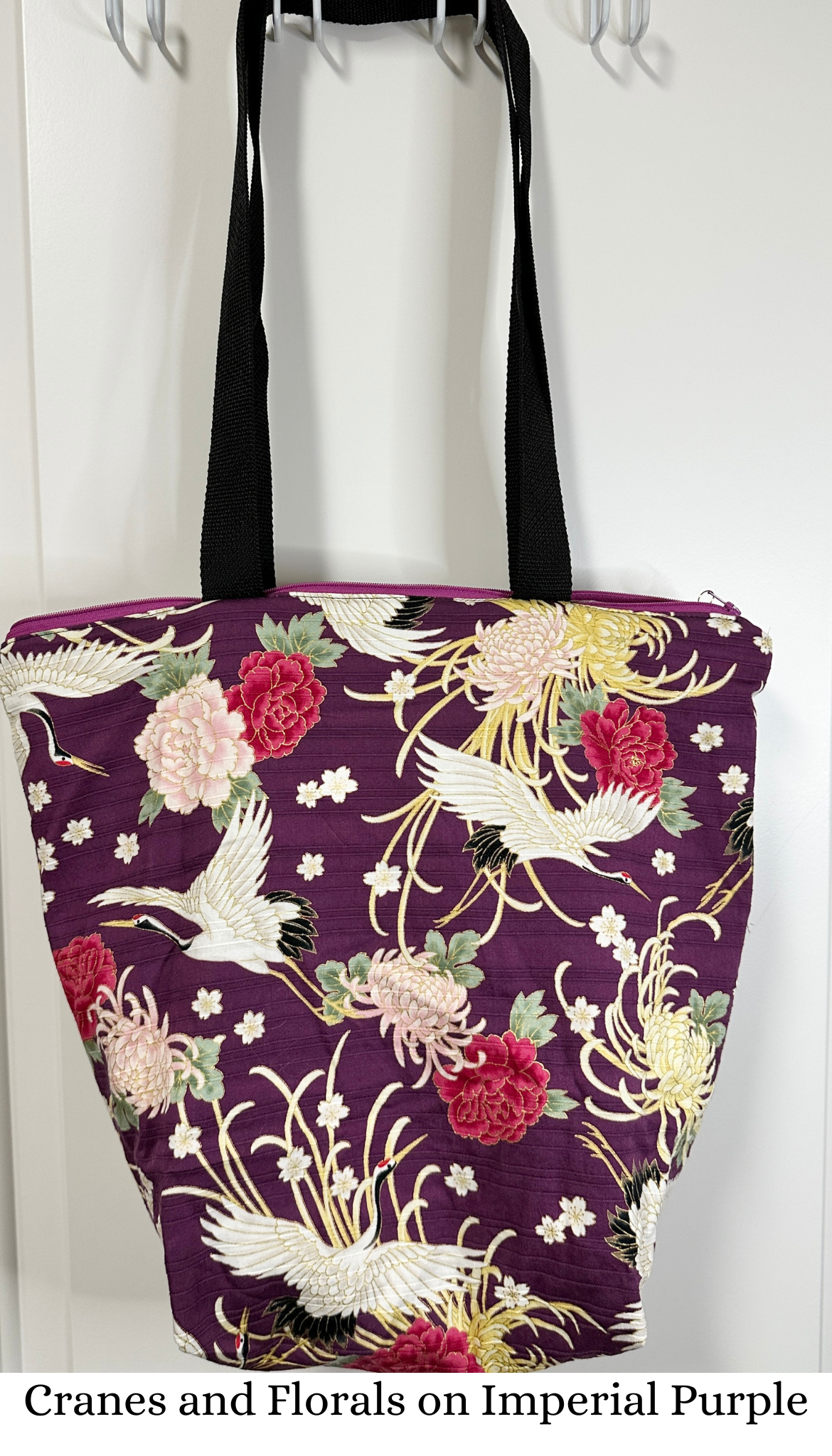 This tote bag has black handles and a purple zipper. The large scale print is pink chrysanthemums and peonies on a background of purple; "flying" over the floral print are Japanese cranes.