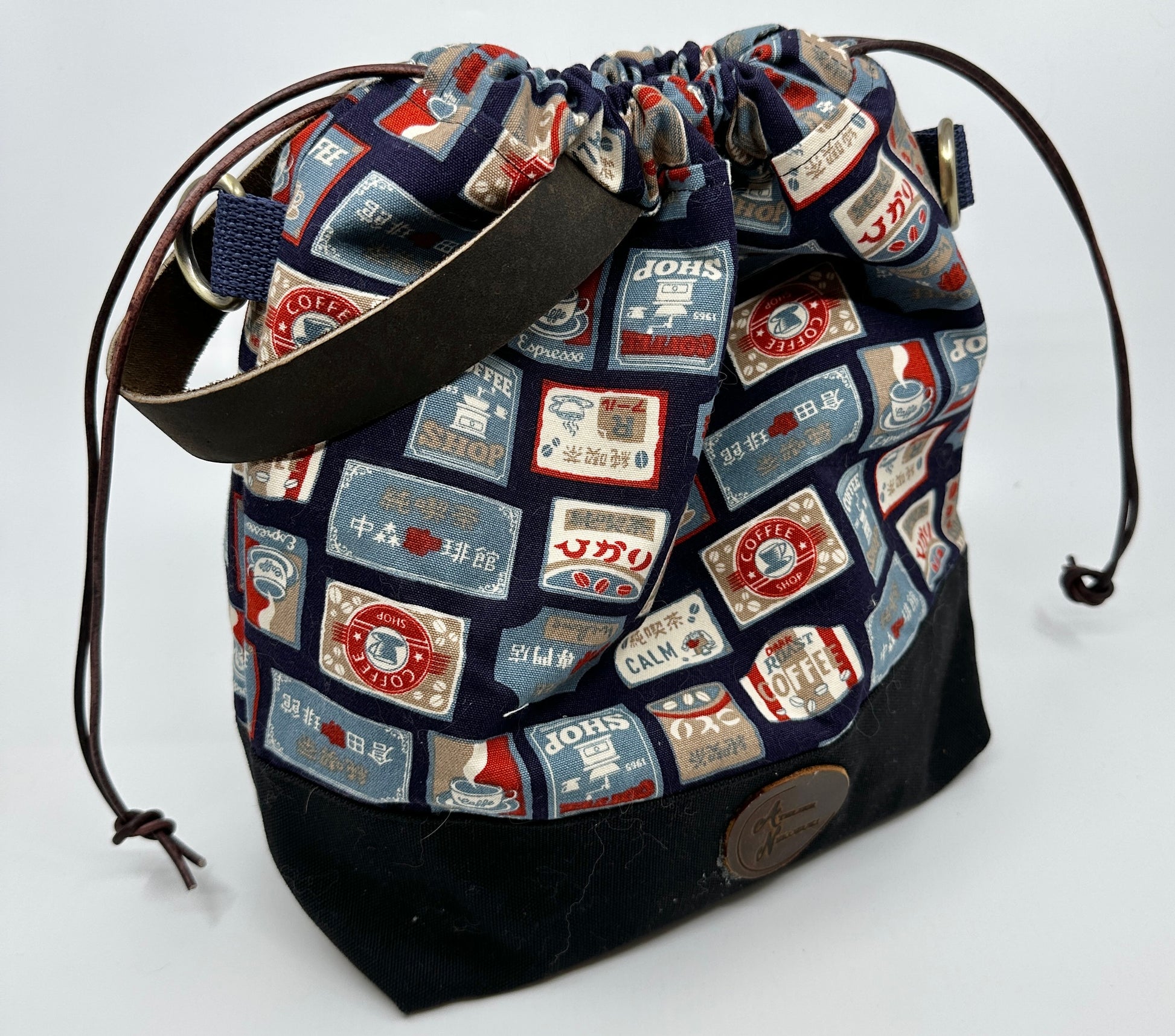 Cinched Kato drawstring bag with navy background. The print is various signs for coffee shops with Japanese text. Leather wristlet handle (sewn into bag) is dark brown; drawstring is dark brown with "distressing"; d rings are bronze. Canvas bottom is black.