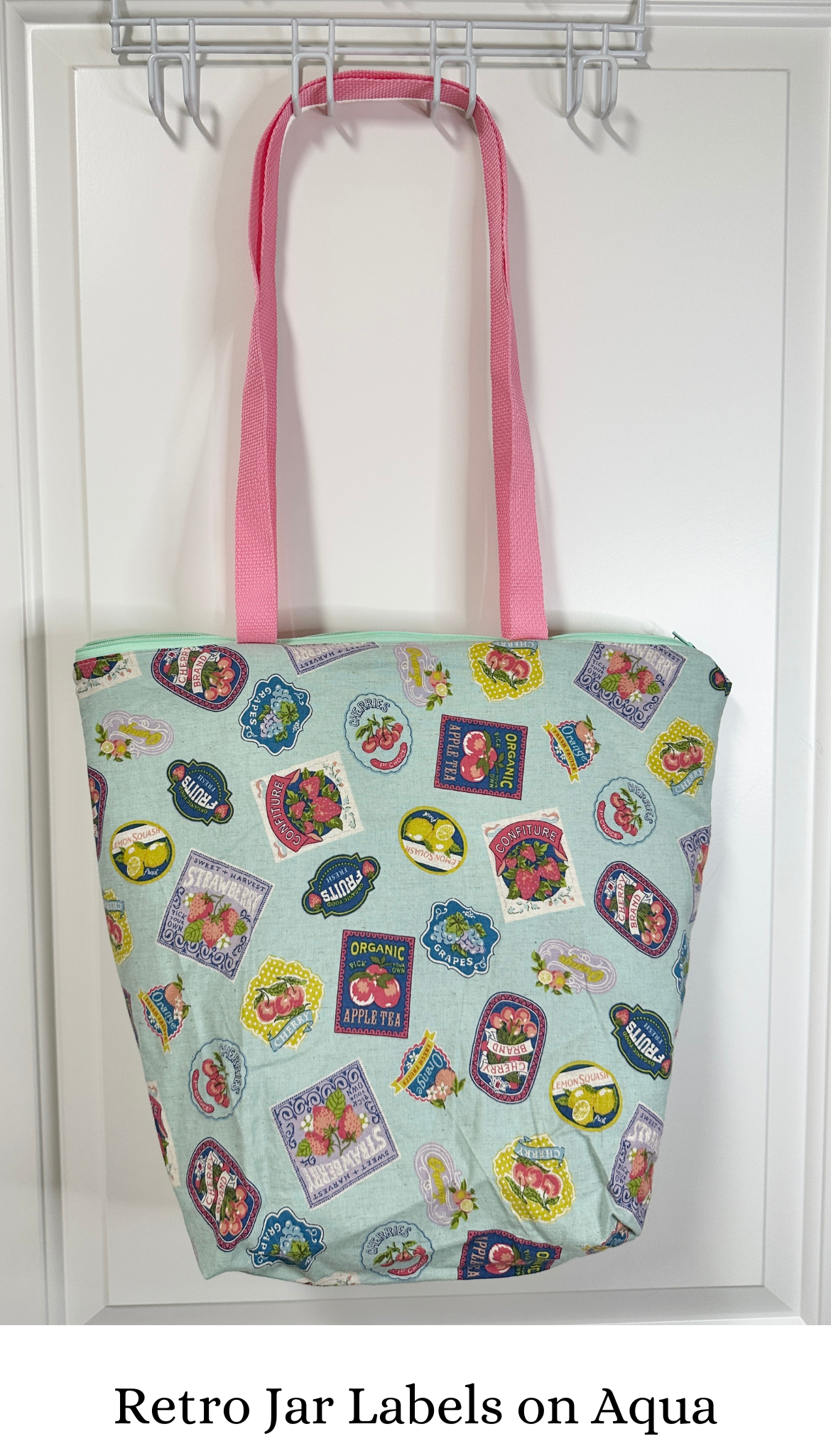 This tote has pink handles and an aqua zipper; the print is various retro styled fruit and jam labels on a background of aqua; the labels are bright against the background. 