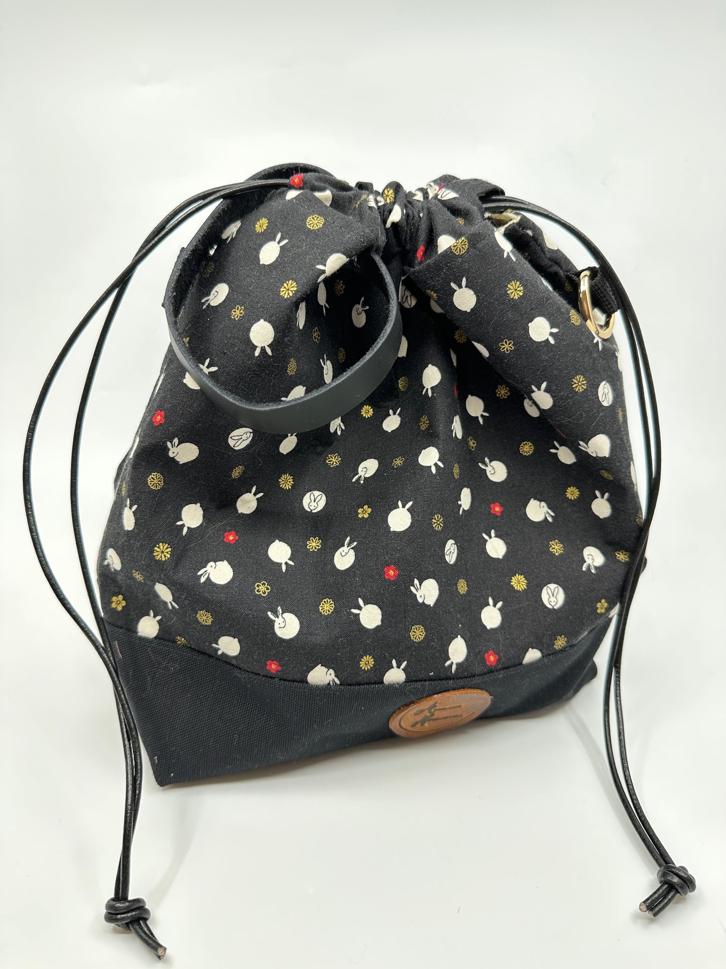 Cinched Kato drawstring bag with black canvas bottom. The Japanese fabric upper on this bag are tiny bunnies that look like polka dots; scattered between bunny are delicate red and shimmering gold flowers. The background of the fabric is black. Drawstring, d ring tabs, and sewn-in leather handle are all black. D rings are gold.