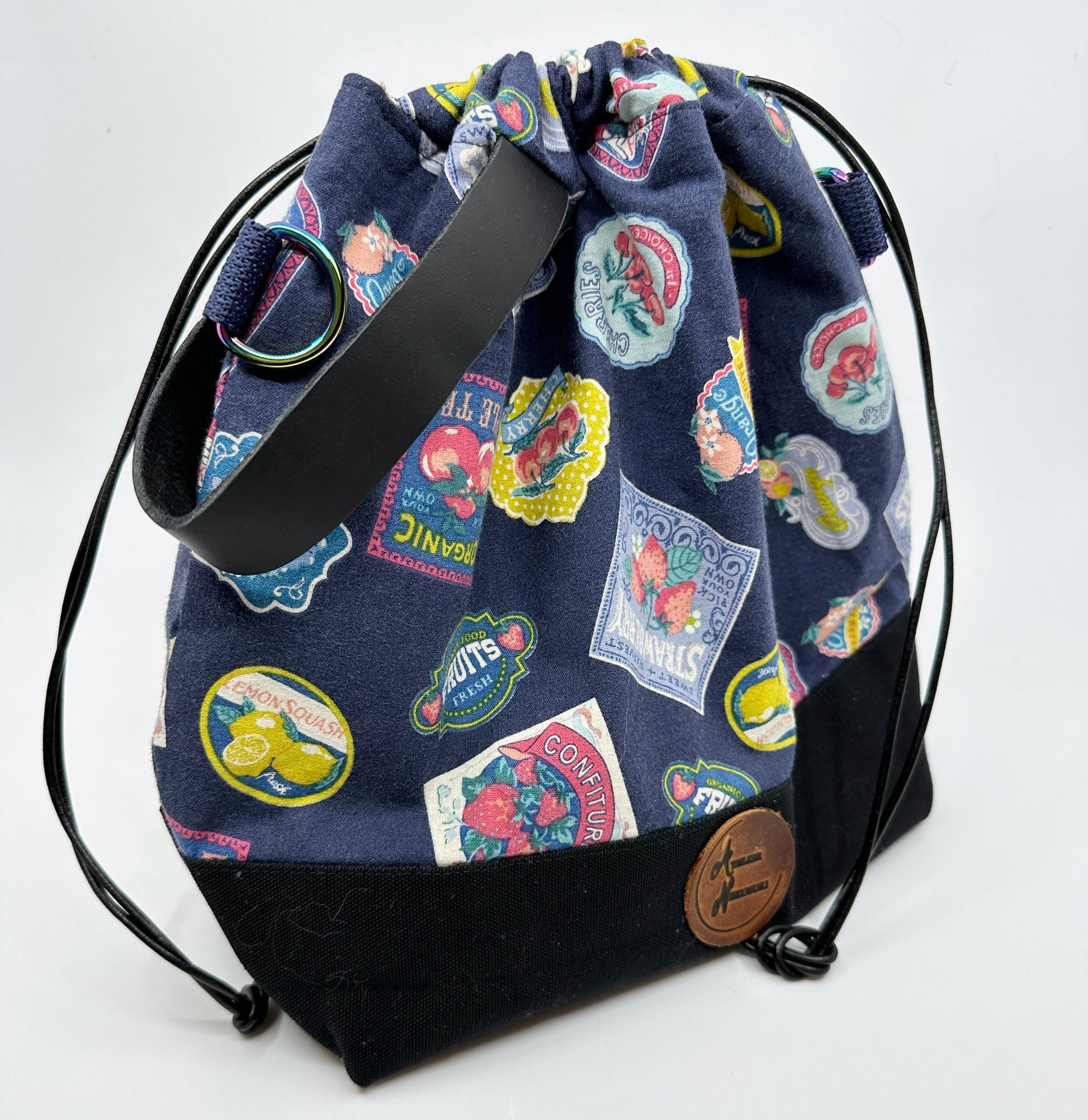 Cinched Kato drawstring bag with retro-styled jam and fruit jar labels on navy background. Canvas bottom is black; leather wristlet handle is black. D-rings for optional cross body strap are iridescent; drawstring is black leather cord.