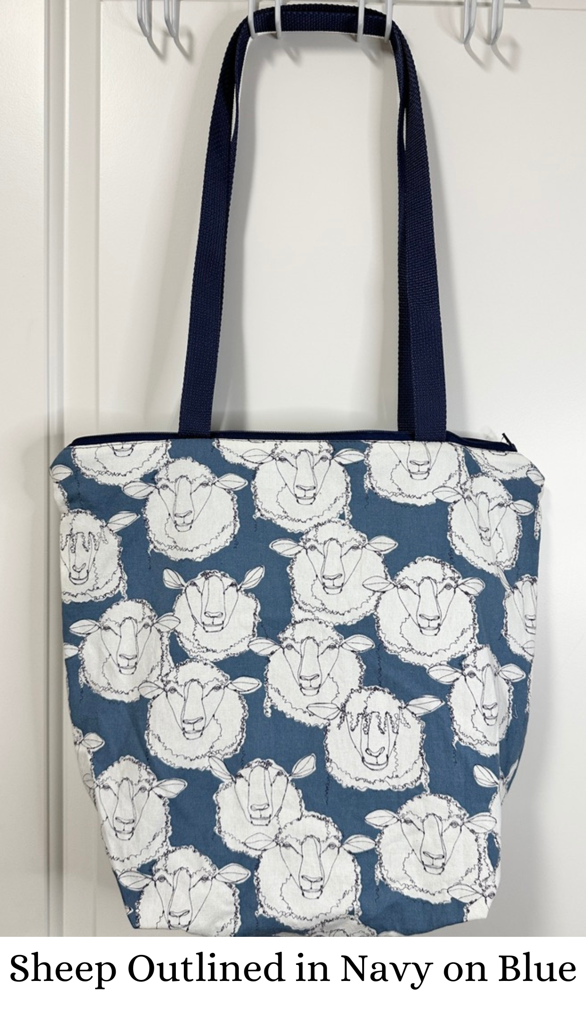 This tote has navy handles and zipper; the print is a large sheep heads on blue. The sheep are outlined in navy/dark blue; there are some shaggy (wool over eye sheep), some fluffy sheep, and some sheep that look like blue faced Leicester.