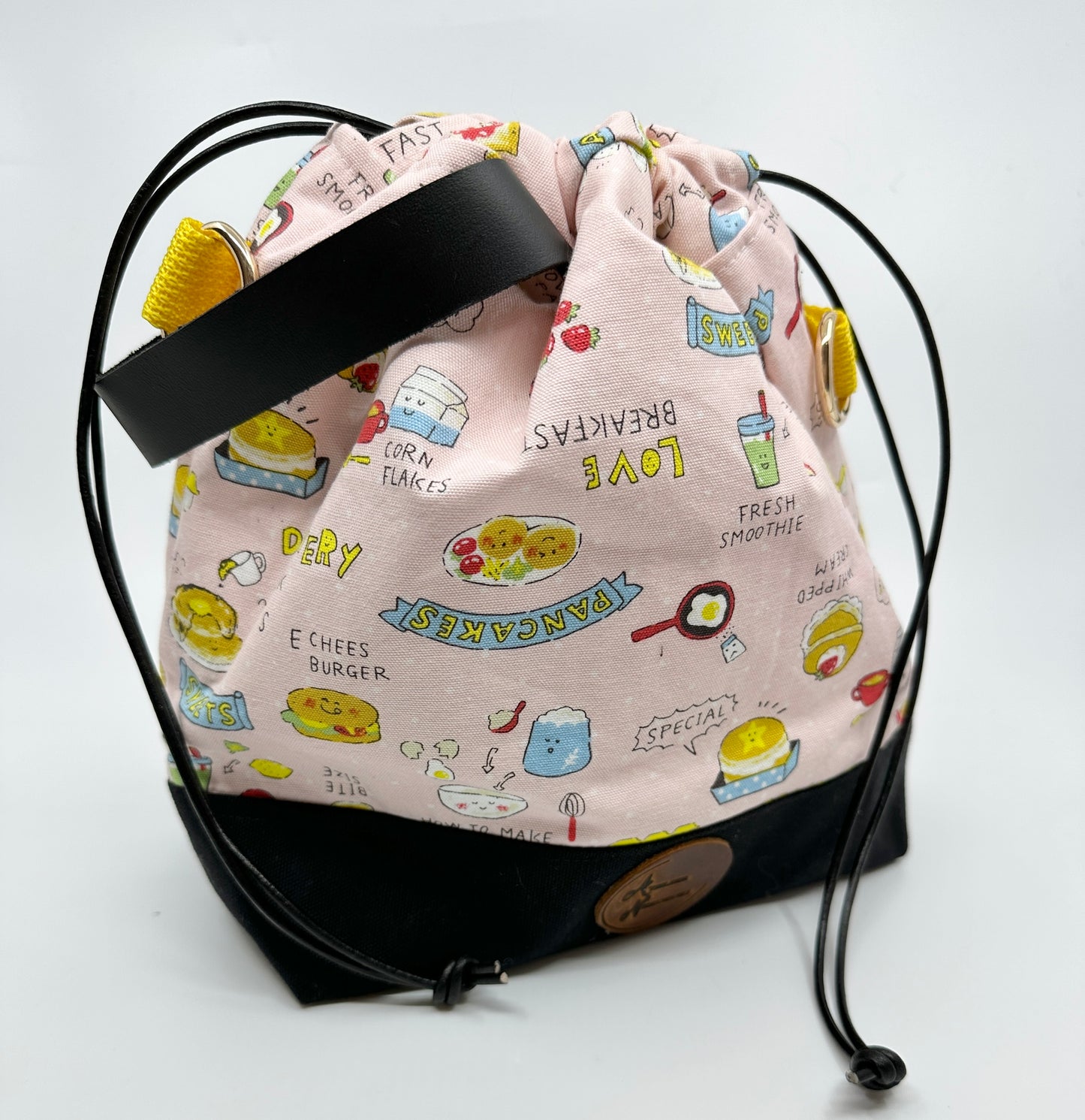 Cinched Kato drawstring bag with black canvas bottom. The Japanese fabric upper is light pink with various breakfast foods (and a random cheeseburger). Sewn in wristlet is black, drawstring is black, d ring tabs are yellow and d rings are gold.