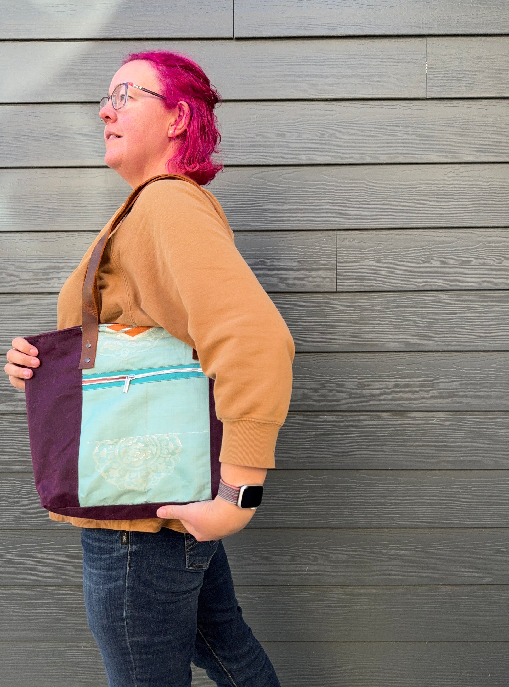 Woman holding tote bag against grey wall; showing off external pocket , which is the same aqua colour with subtle patterning at bottom. 