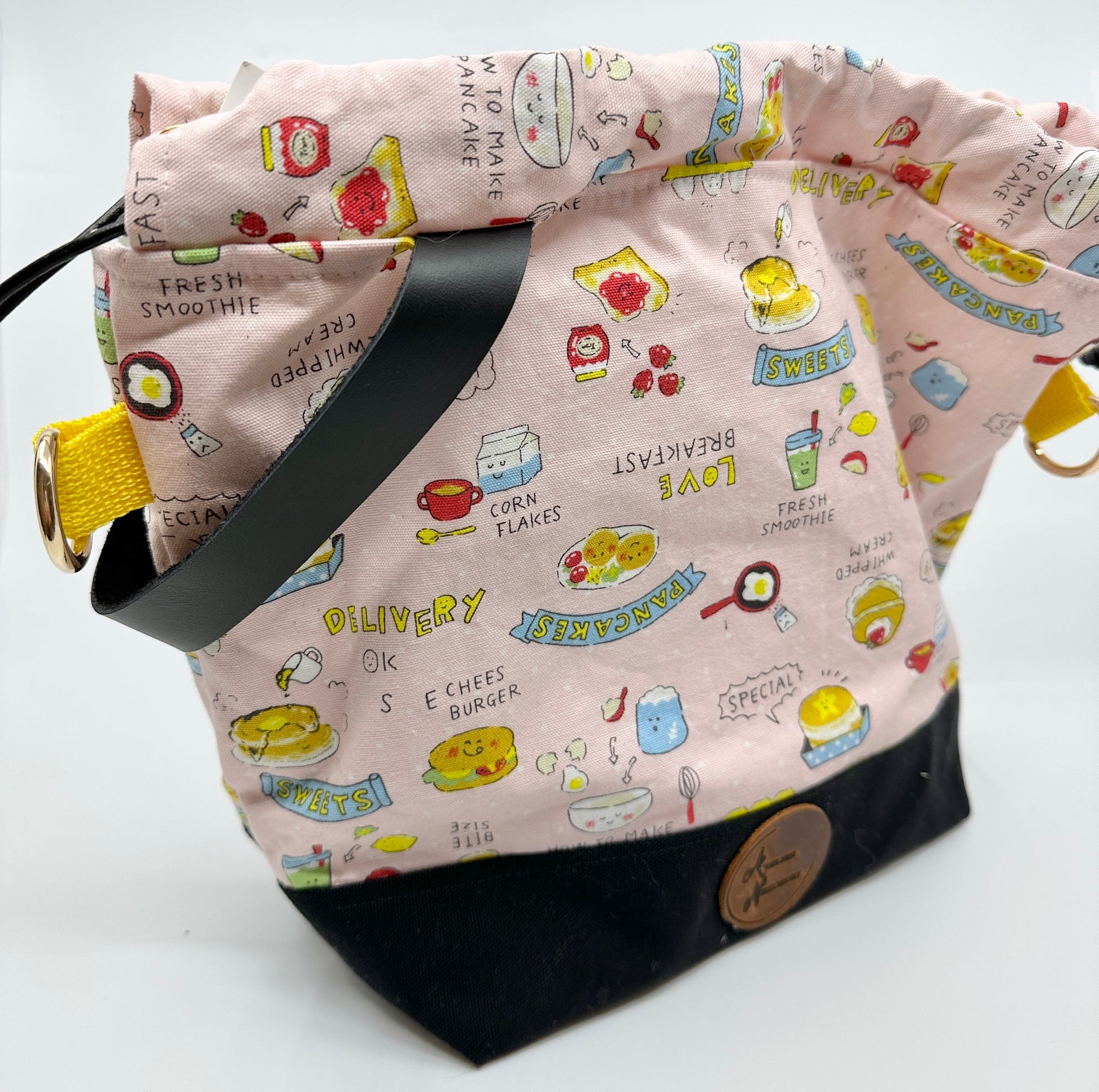 Open Kato drawstring bag with black canvas bottom. The Japanese fabric upper is light pink with various breakfast foods (and a random cheeseburger). Sewn in wristlet is black, drawstring is black, d ring tabs are yellow and d rings are gold.