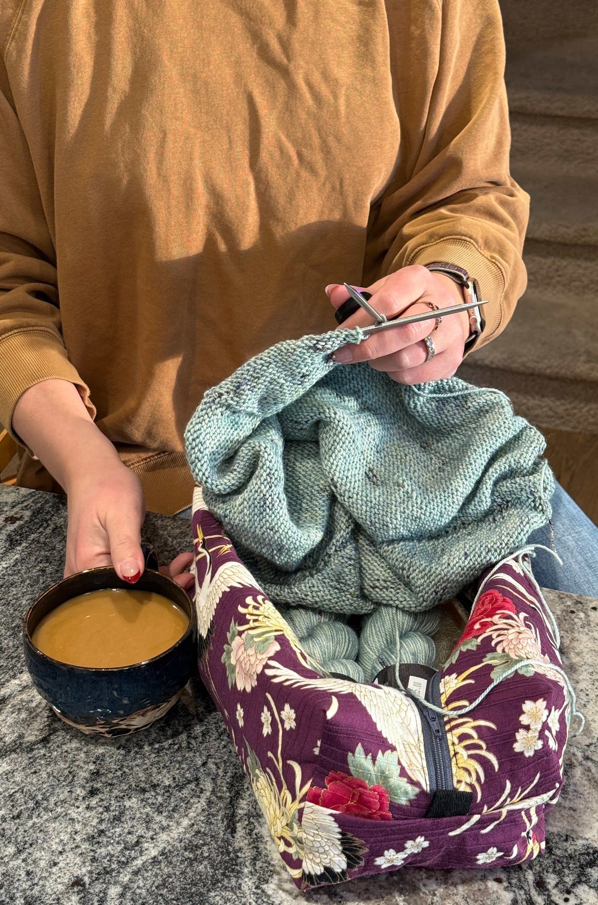 Person knitting with a green yarn and holding a cup of coffee, cranes on purple project bag in front