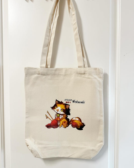 Off-white large tote bag has a centred image of the Atelier Nekozuki logo - a calico cat in white, orange, and brown. There is a red ball of yarn skewered with knitting needles to the left and to the right above the cat's ear, there is the company name "Atelier Nekozuki" in black, with Nekozuki also in red Japanese kanji & hiragana.