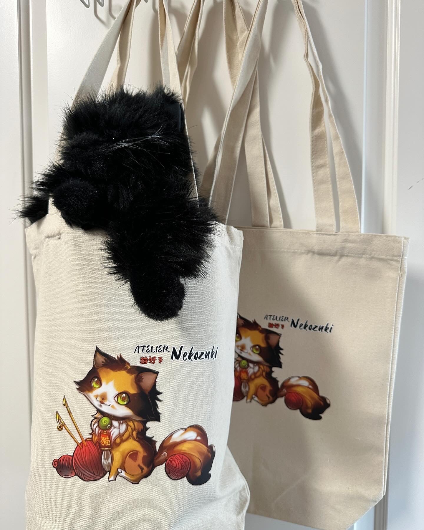 Off-white large tote bag has a centred image of the Atelier Nekozuki logo - a calico cat in white, orange, and brown. There is a red ball of yarn skewered with knitting needles to the left and to the right above the cat's ear, there is the company name "Atelier Nekozuki" in black, with Nekozuki also in red Japanese kanji & hiragana. In the bag on the left is a handmade stuffed animal that is a black cat (also from Japan, his name is Chachamaru.)