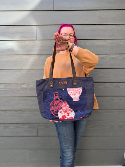 Woman holding a navy tote bag with Japanese vases elements against a gray wall.
