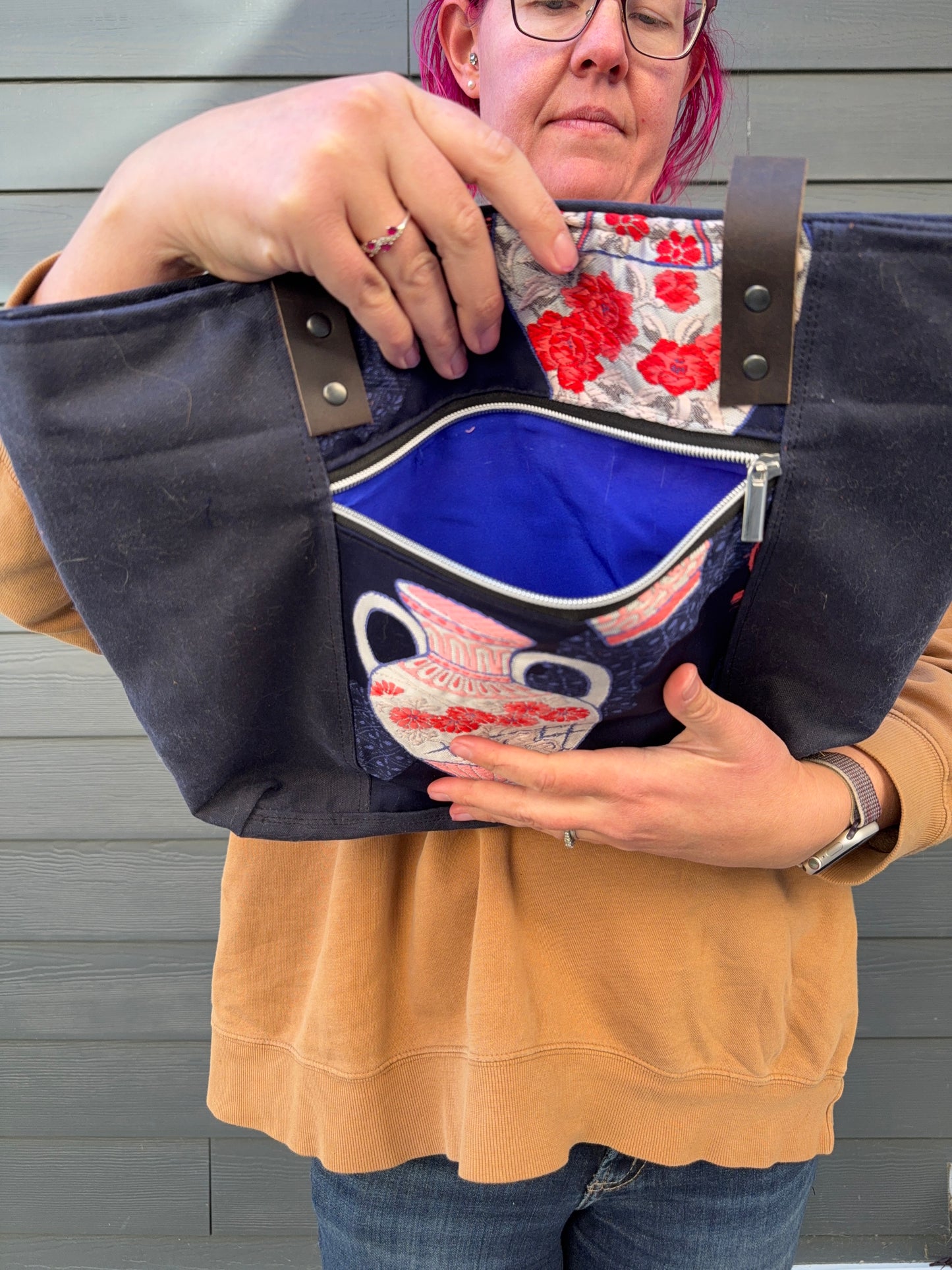 Woman holding a navy tote bag with Japanese vase elements against a gray wall. Showing bright blue pocket lining for exterior zip pocket.