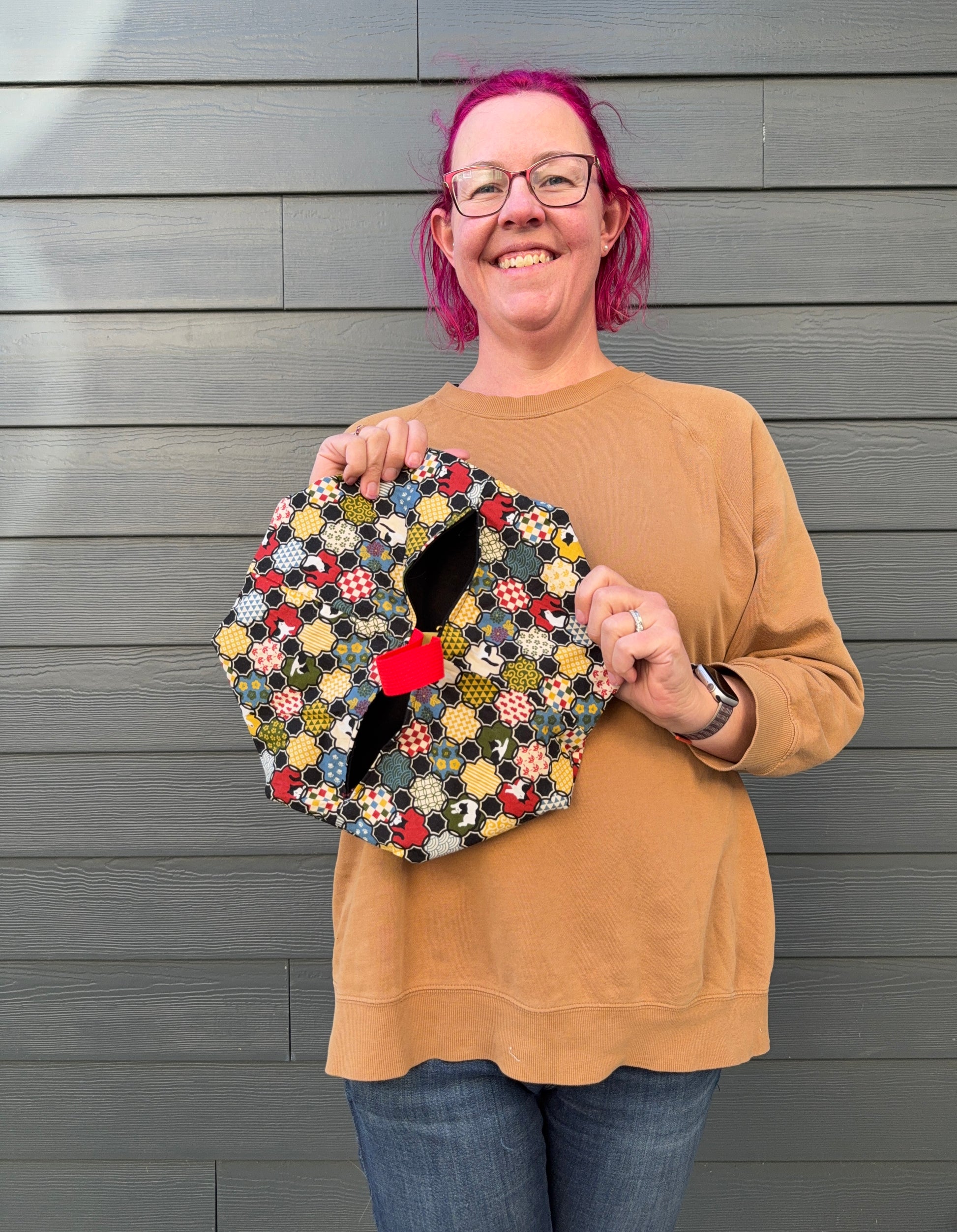 Person holding a colourful quilt patterned bag against a grey wooden wall. Pattern is Tuxedo cats and Japanese motifs in blue, red, green, and gold.