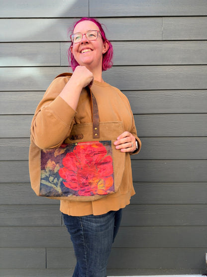 Woman holding a tote bag against a grey wall; detail is a large poppy against navy. The poppy is framed by tan waxed canvas.