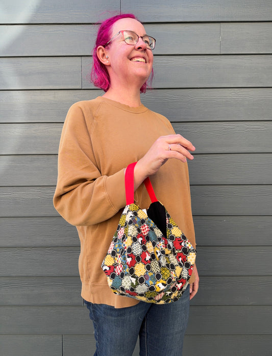 Person holding a cat and Japanese patterned  handbag  in blue, red, green, and gold colours with a red strap against a grey wooden wall.