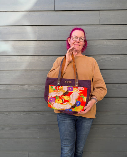 Person holding a colourful bag with cranes and large florals on orange against a grey wall. Brown leather and gold accents.